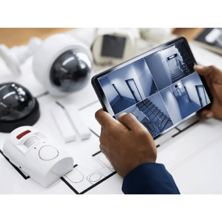 Hands hold a tablet showing a grid of security camera feeds over architectural blueprints on a desk with cameras and a smoke detector nearby.