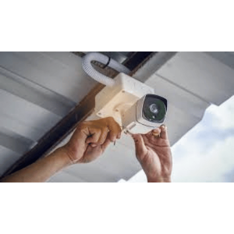 Technician installing an outdoor security camera on a wooden mounting block under a metal overhang.